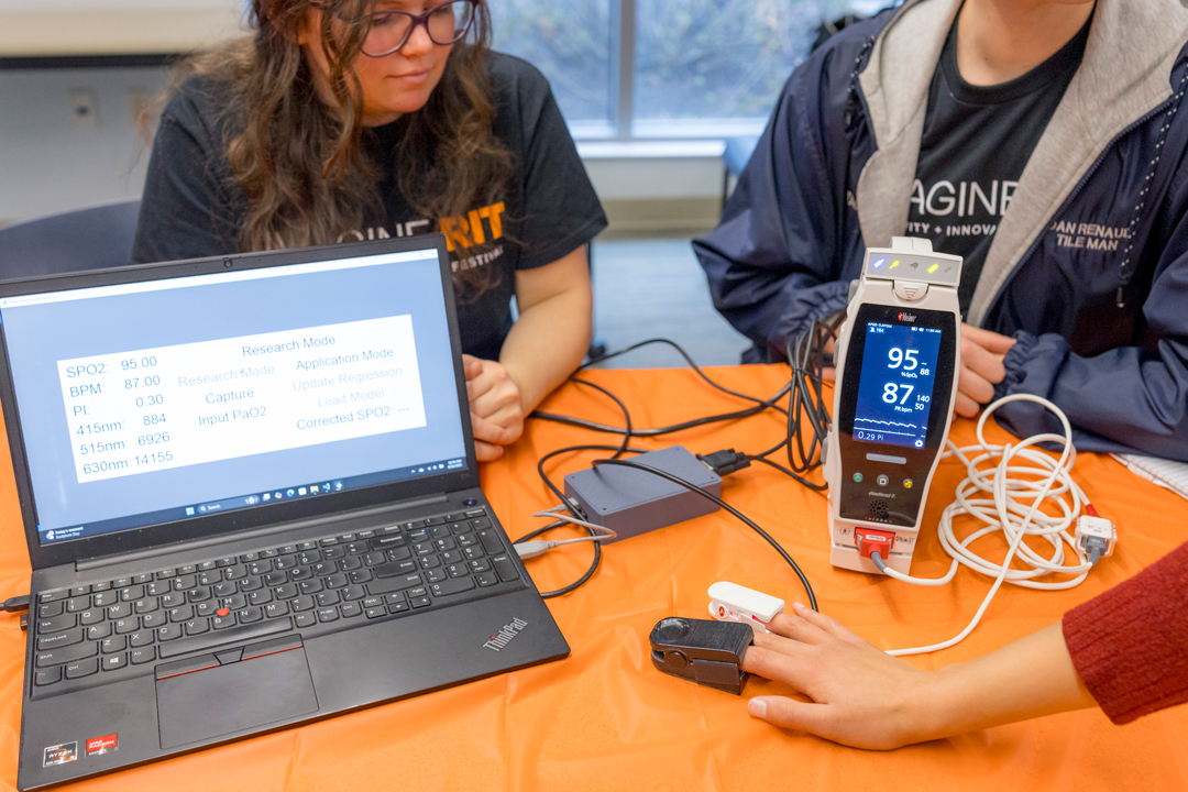 Two people sit at a desk with an open laptop and oximeter device.